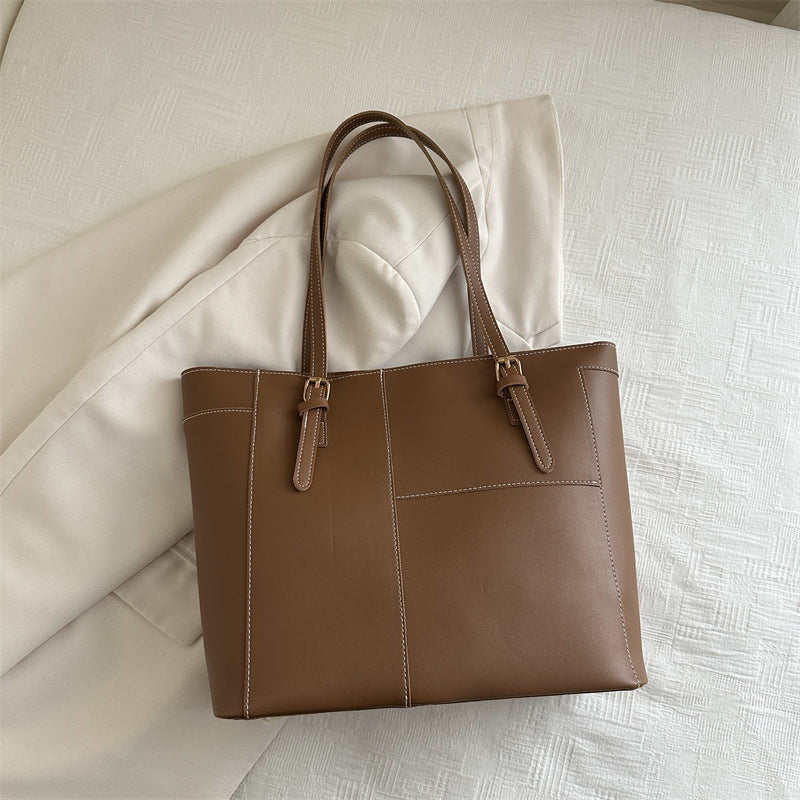 Brown large shoulder bag with double handles and buckle details placed on beige fabric