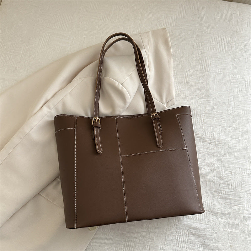Brown large shoulder bag with double handles and stitched detailing on beige fabric background