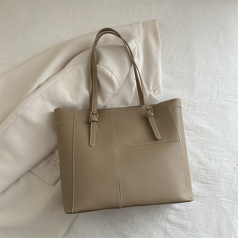 Beige large shoulder bag with adjustable straps placed on a cream jacket on white textured surface