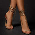Gold ankle strap stiletto sandals with wrap-around ties on model feet by Sheila Merchandise